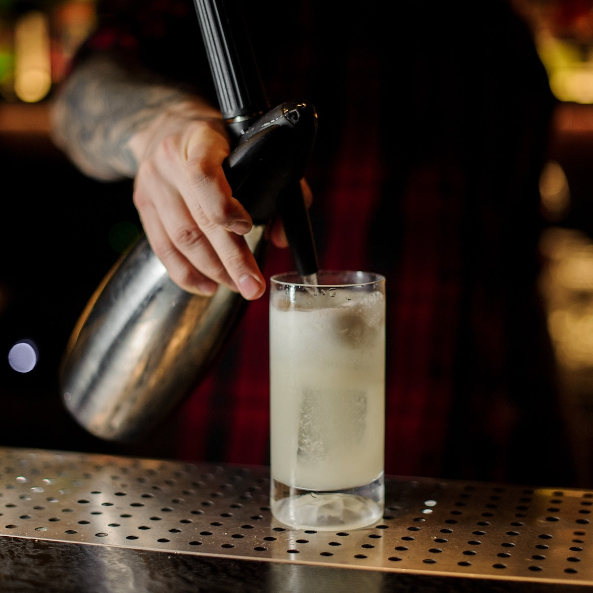 A bartender in a plaid shirt fills a tall glass with ice cubes, using a silver soda siphon to add Top Hat Provisions Top Hat Extra Spicy Ginger Beer Syrup & Batching Concentrate for bold flavor. The glass rests on a perforated bar counter, warmly illuminated by the softly blurred ambient lighting.