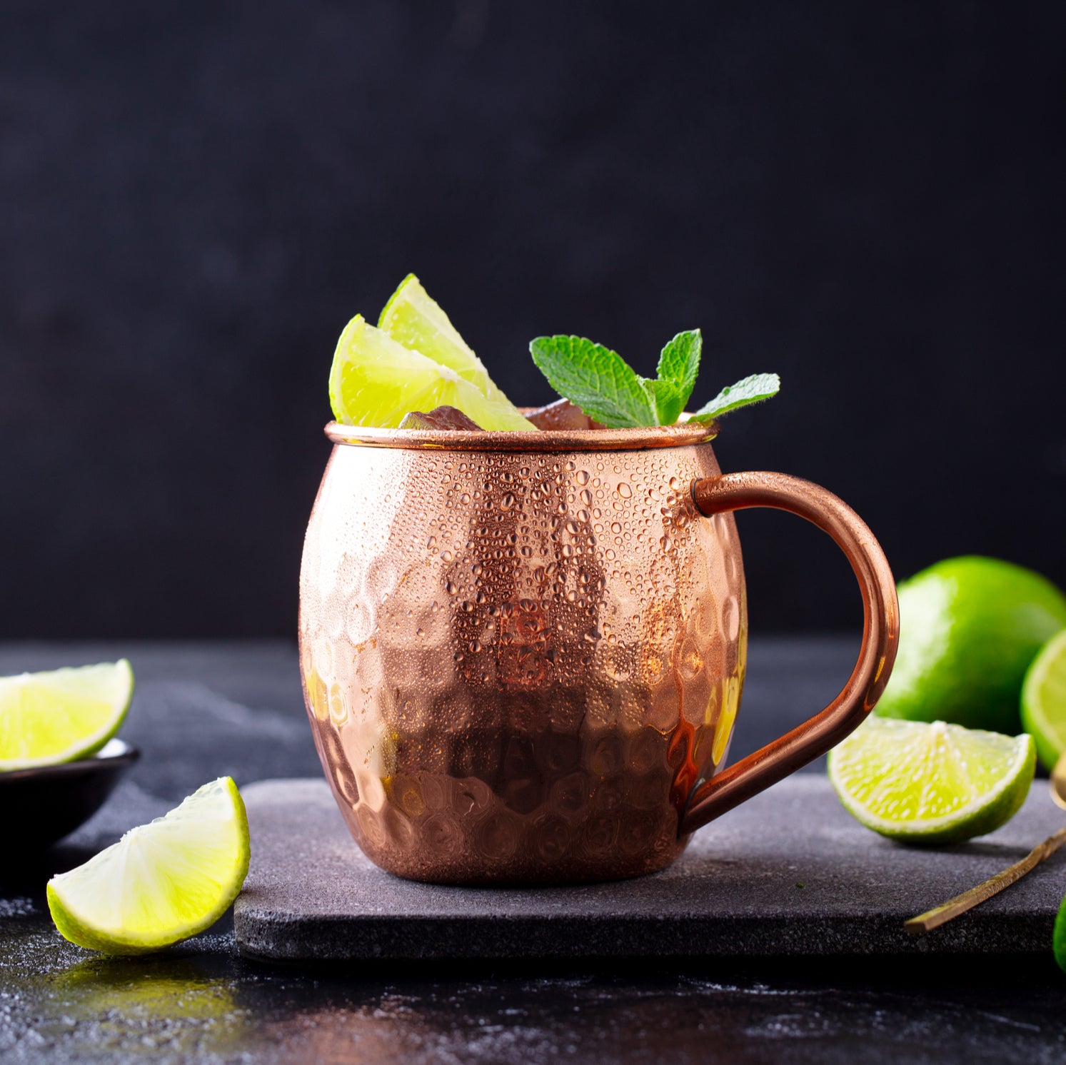 A copper mug loaded with a Moscow Mule is adorned with lime wedges and mint, sitting on dark slate. Extra lime hints at the zesty craft from Top Hat Extra Spicy Ginger Beer Soda Syrup by Top Hat Provisions in this refreshing cocktail.