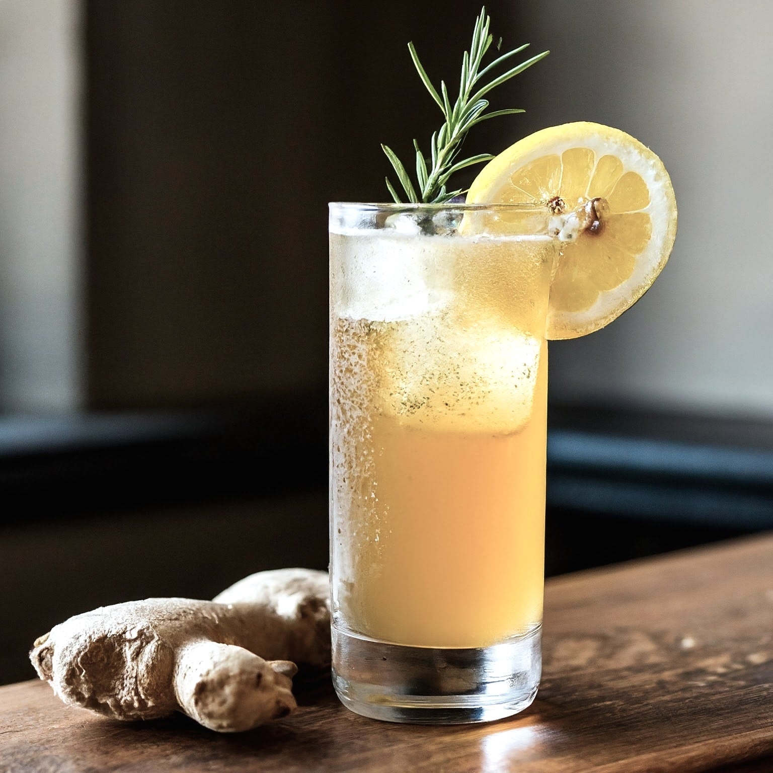 On a wooden surface stands a tall glass filled with an iced beverage made from Top Hat Provisions Extra Spicy Ginger Beer BIB Soda Syrup, garnished with lemon and rosemary. Nearby lie two ginger root pieces, enhancing the fresh look.