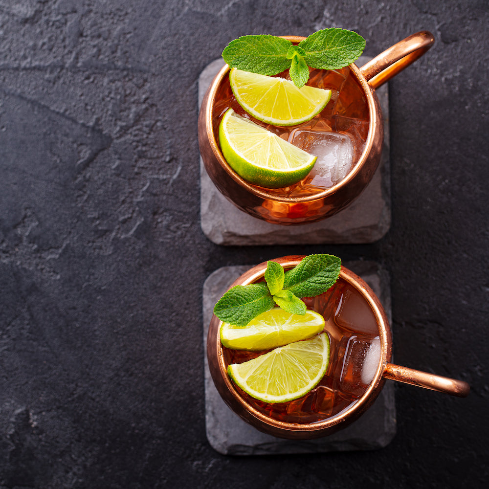 Two copper mugs of iced Moscow Mules, enhanced with Top Hat Provisions Extra Spicy Ginger Beer Syrup, are garnished with lime wedges and fresh mint, resting on square slate coasters against a dark textured background.