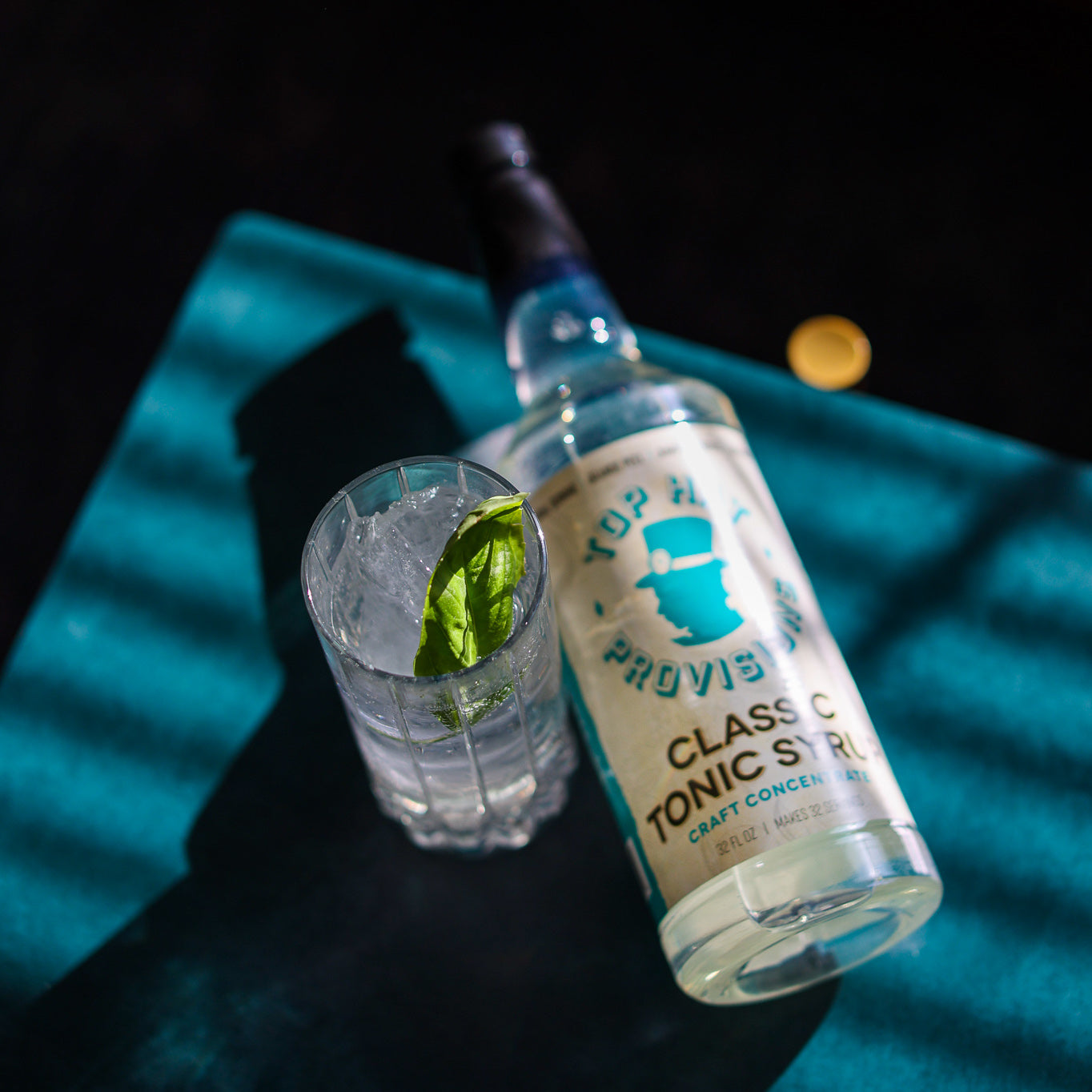 Classic tonic bottle on side in louver light with G&T and basil, square crop