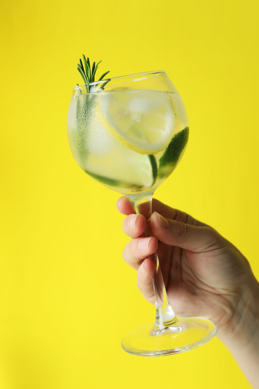 Light cocktail in goblet held high with yellow backdrop, rosemary sprig, and lemon