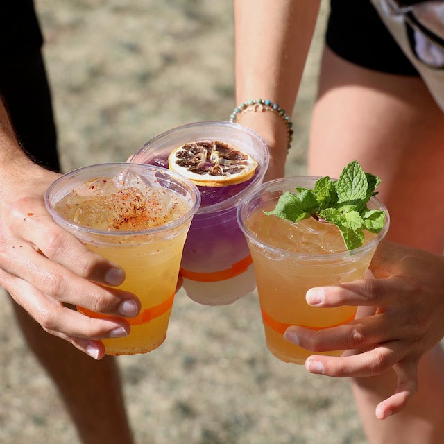 Three people with clear cups of colorful cocktails: one garnished with dried citrus, the second topped with mint, and the third looking like an extra spicy mule made with Top Hat Provisions Extra Spicy Ginger Beer Syrup. The backdrop is a blur of grass.